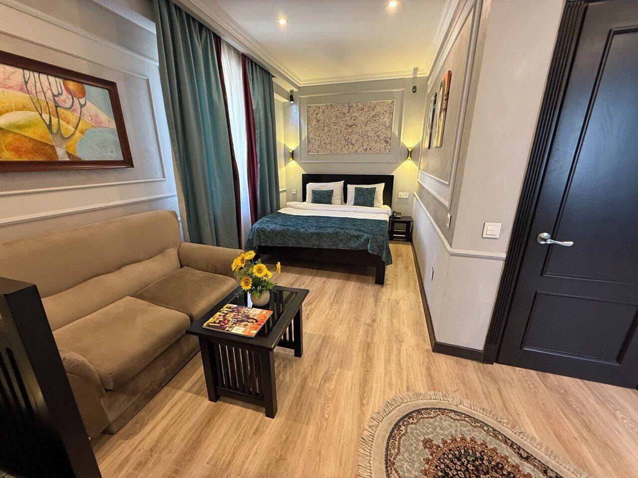 A hotel room with a sofa, a bed with a green bedspread, a small table with a vase of sunflowers, and a large painting on the wall.