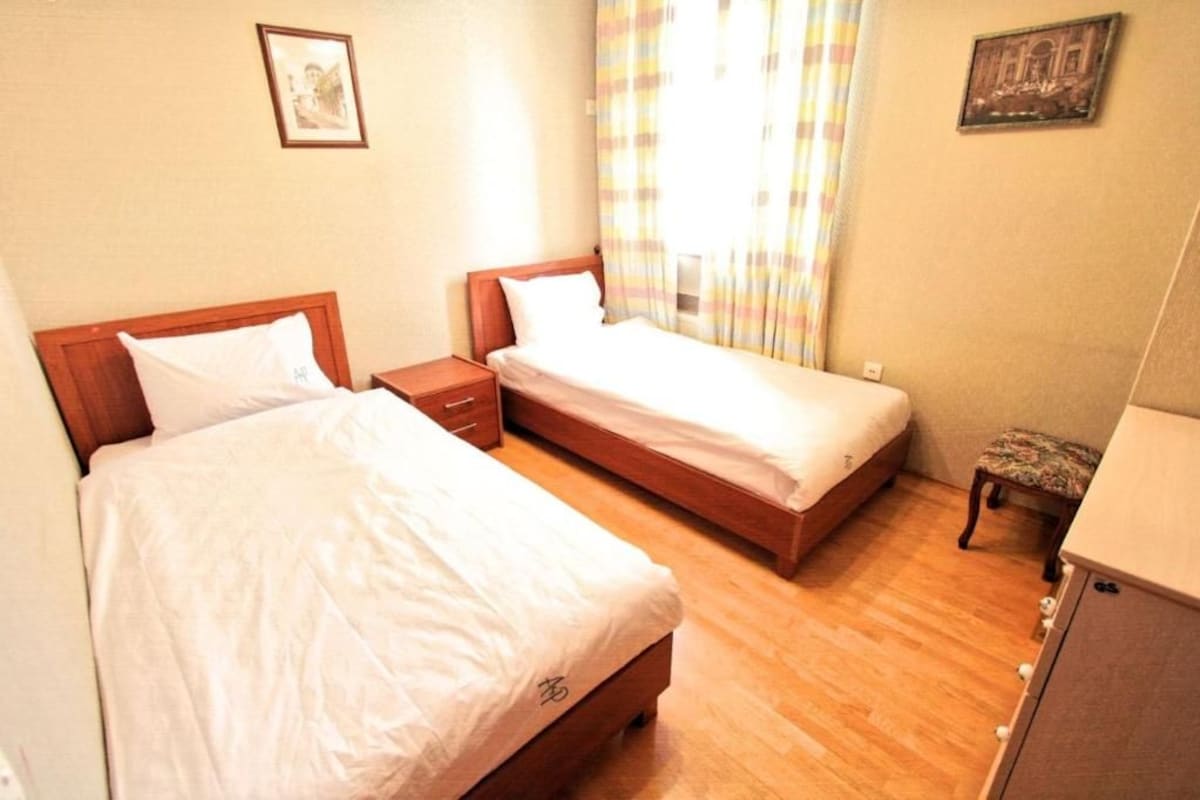 Hotellihuone, jossa on kaksi erillistä sänkyä, puinen yöpöytä, pieni pöytä ja penkki.