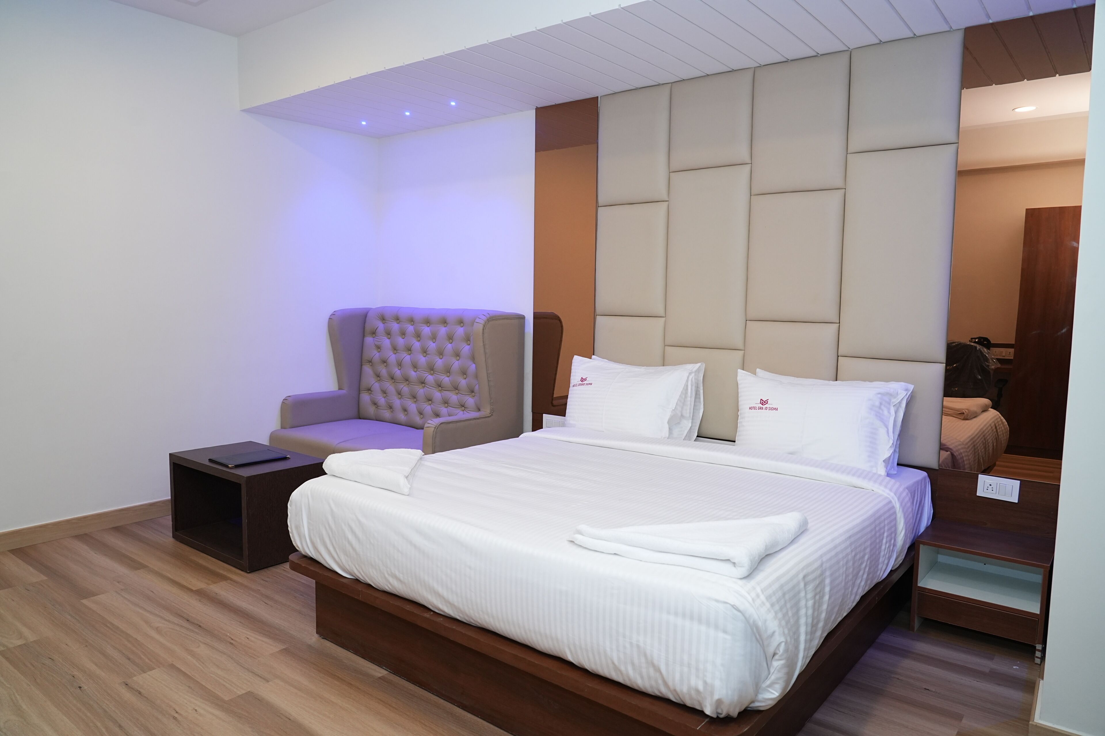 A modern hotel room with a large bed, a tufted armchair, a small bedside table, and a wooden headboard.