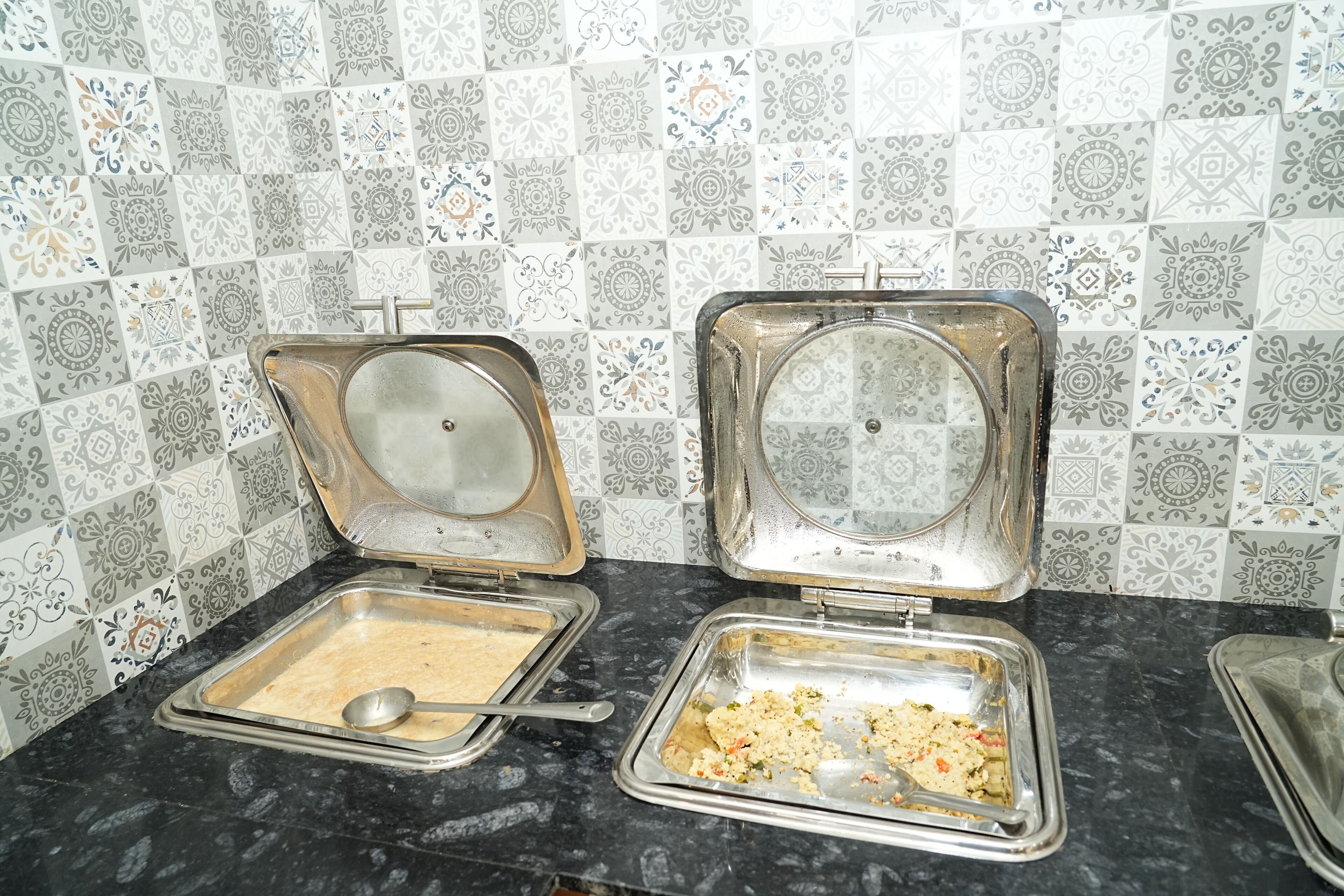 A kitchen area with two open metal trays on a black countertop, one containing a creamy substance and the other with a mixture of grains and vegetables.