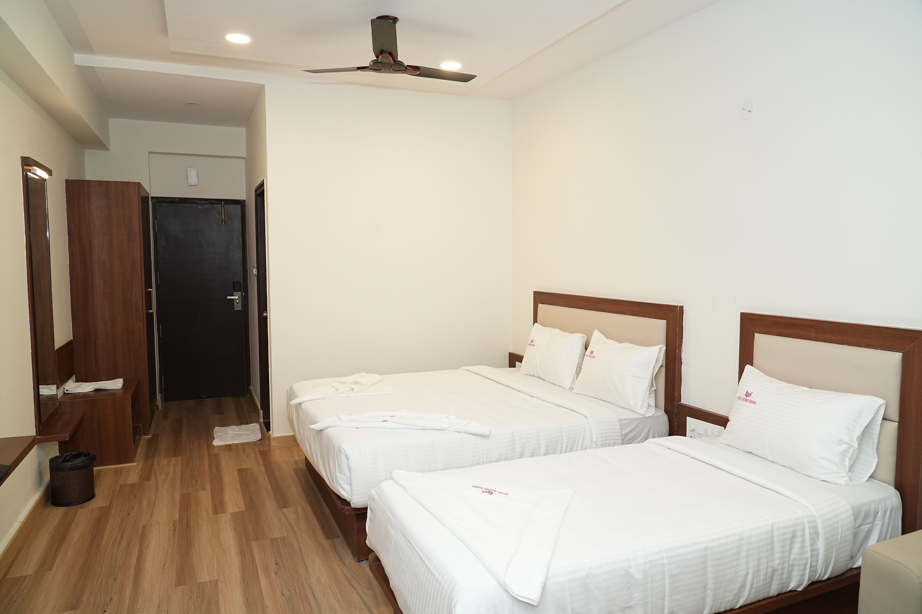 Una habitación de hotel con dos camas individuales, cabeceros de madera, ropa de cama blanca, un ventilador de techo y piso de madera.