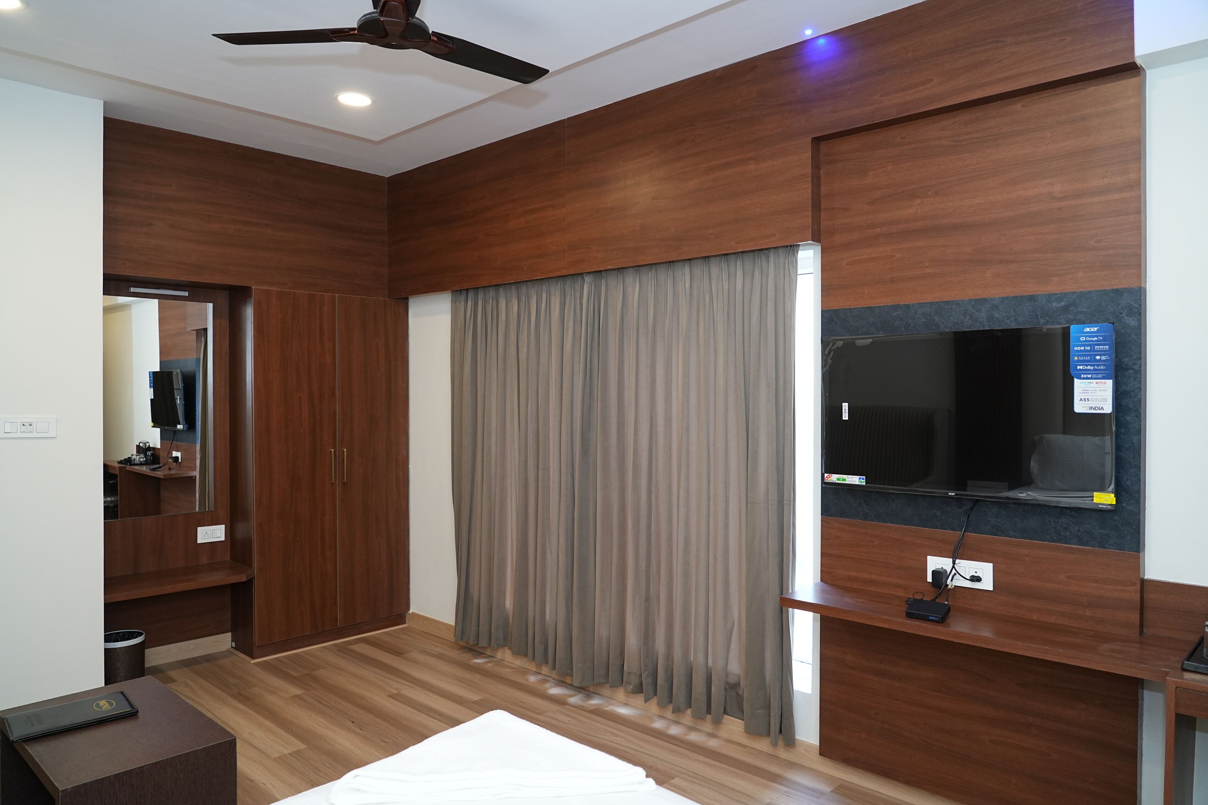 A modern hotel room with a wooden floor, a bed, a TV mounted on the wall, a ceiling fan, and a large window with curtains.