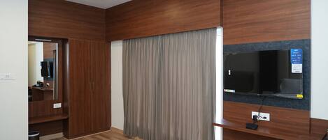 A modern hotel room with a wooden floor, a bed, a TV mounted on the wall, a ceiling fan, and a large window with curtains.