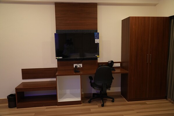 A room with a wooden desk, a black office chair, a television mounted on the wall, and a wardrobe.