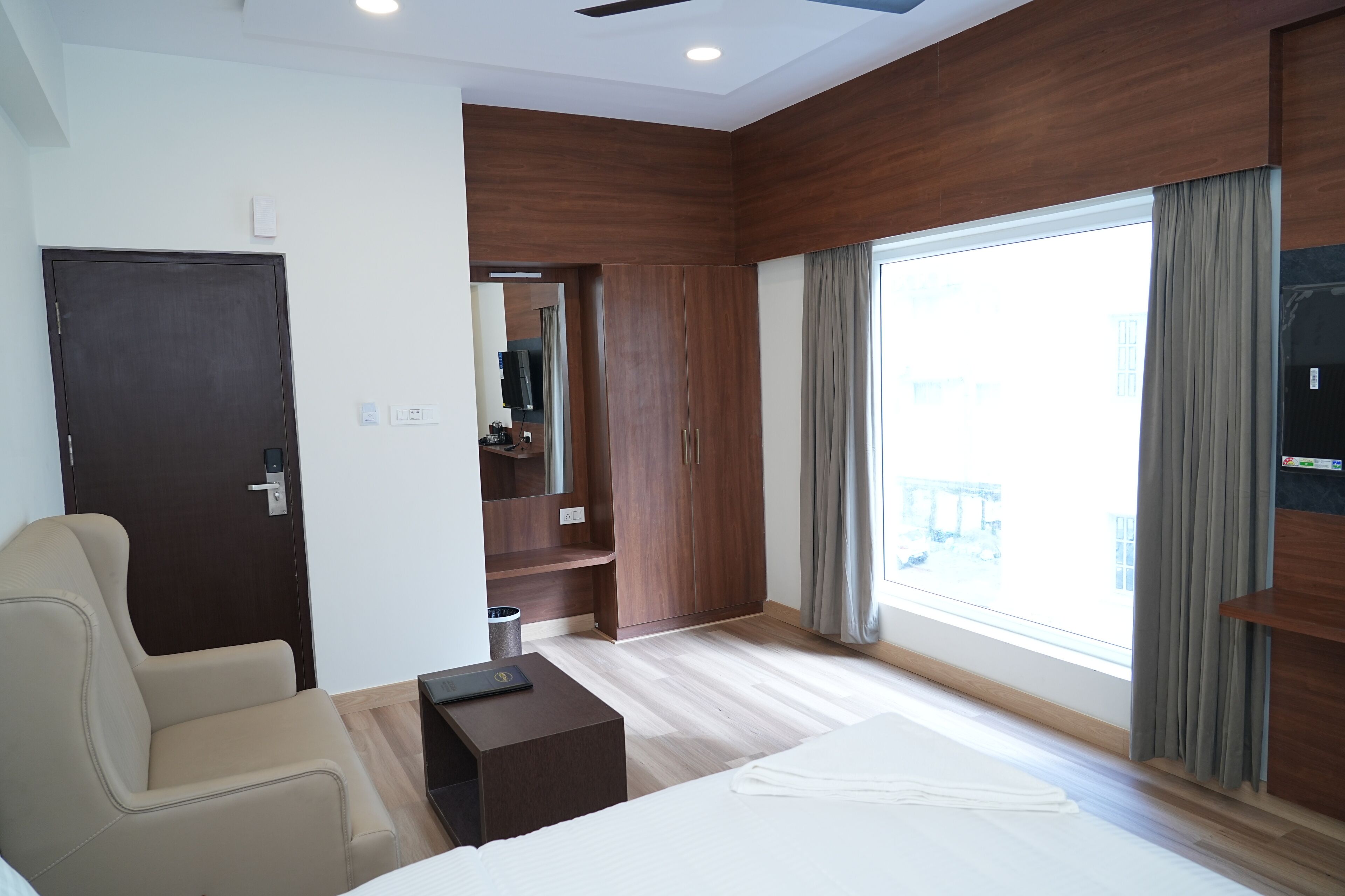 A modern hotel room with a large bed, a seating area, a wardrobe, and a view of the outside.