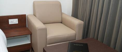 A hotel room with a beige armchair, a wooden nightstand with a power outlet, and a small table with a Dell laptop.