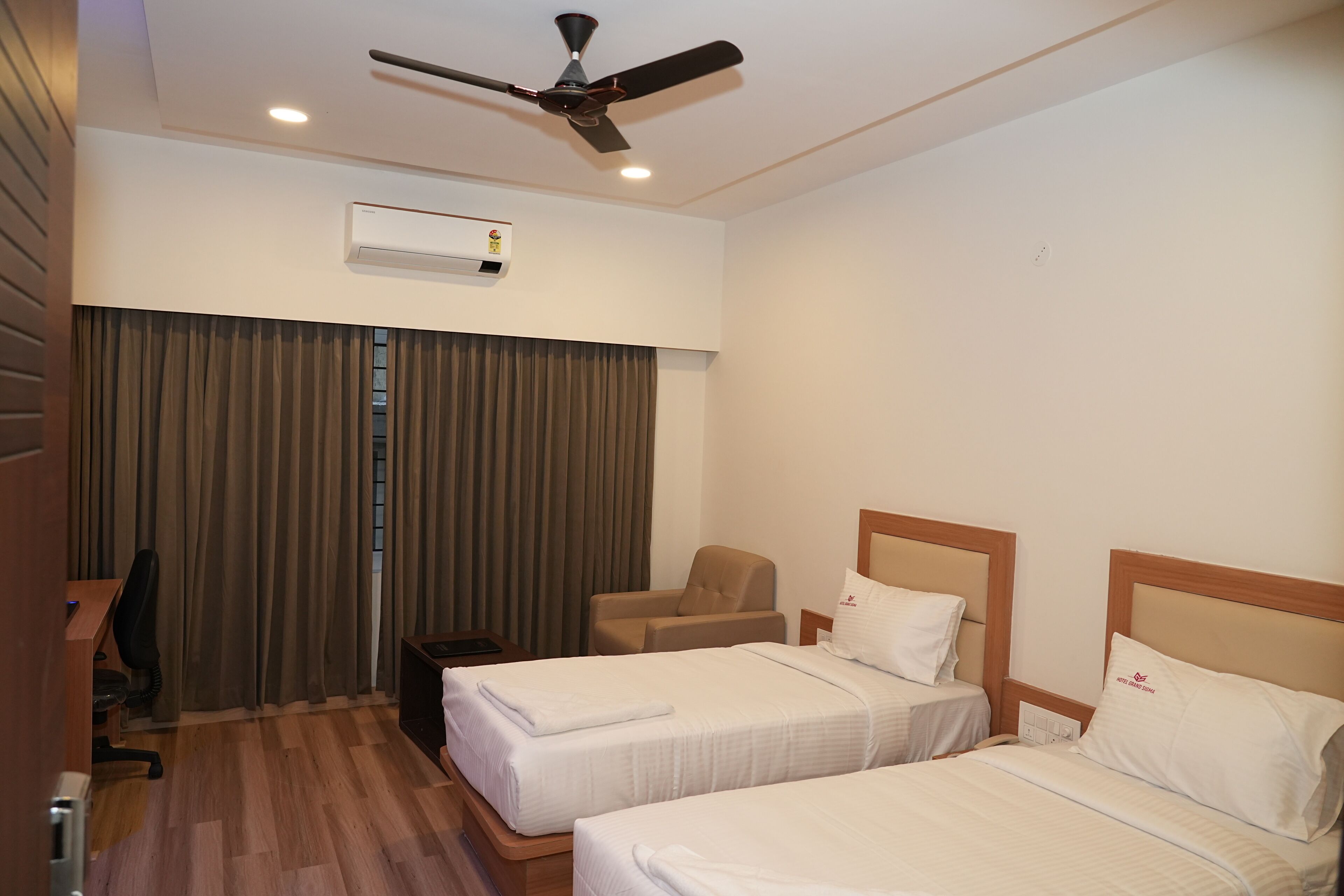 A hotel room with two beds, a desk, a chair, an air conditioning unit, and a ceiling fan.