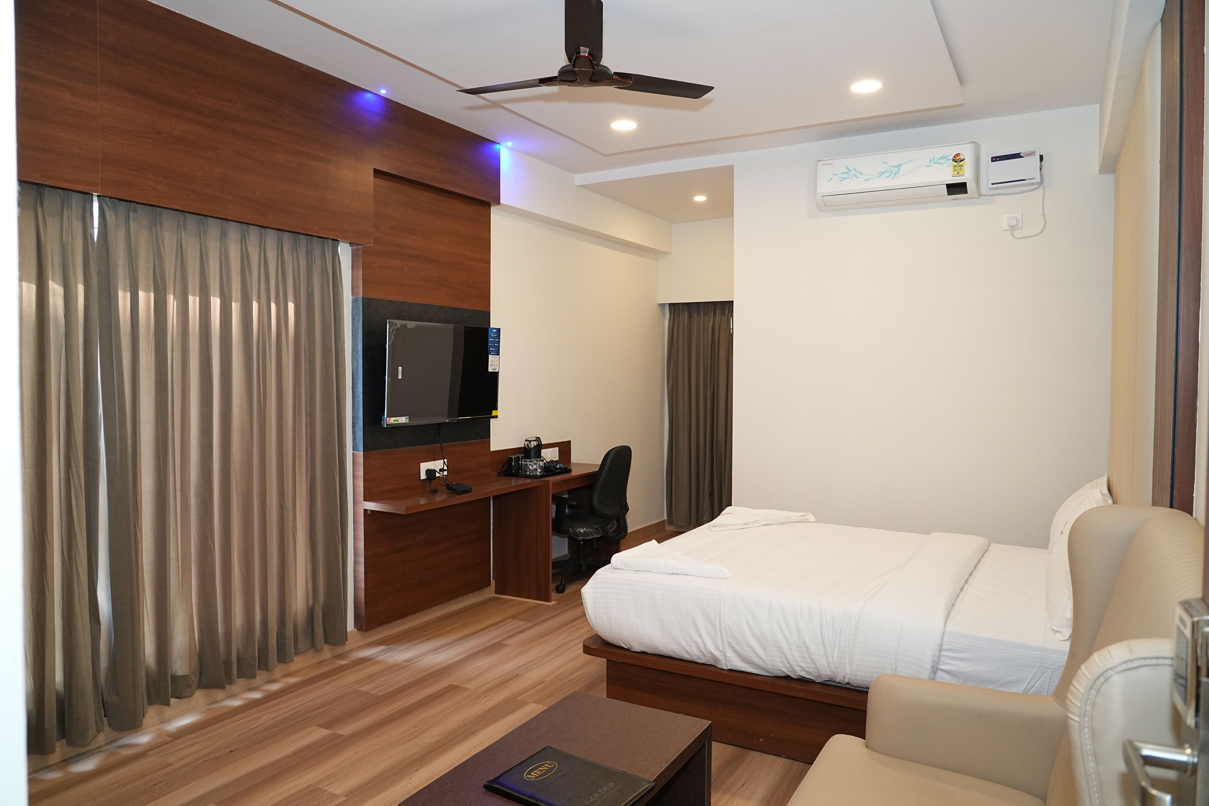 A modern hotel room with a bed, a desk, a chair, a television, and an air conditioning unit.