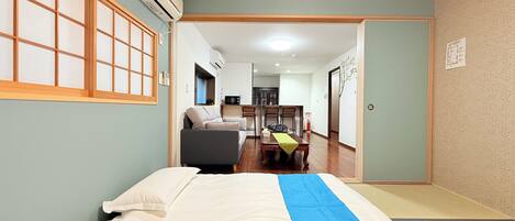 A room with a bed, a sofa, a dining table with chairs, and a kitchen area in the background.
