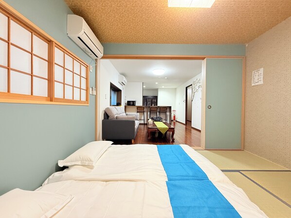 A room with a bed, a sofa, a dining table with chairs, and a kitchen area in the background.
