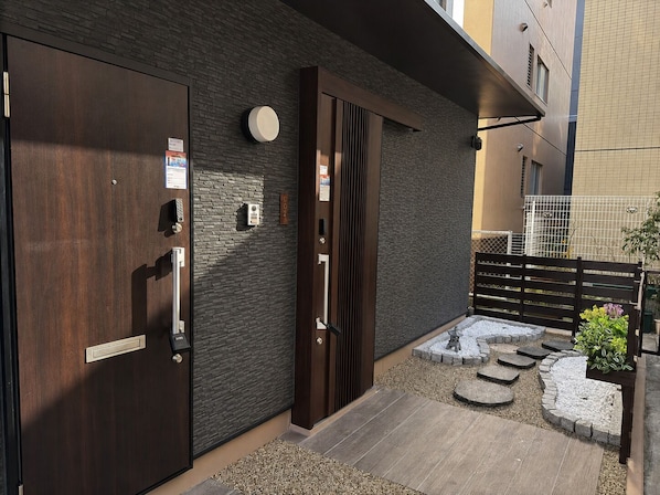 A modern entrance with a wooden door, a keypad, and a small outdoor seating area with steps and a garden.