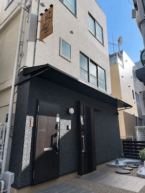 Un bâtiment moderne doté d’un toit plat, d’une enseigne en bois ornée de caractères japonais et d’une porte équipée d’un clavier pour l’ouverture à code.