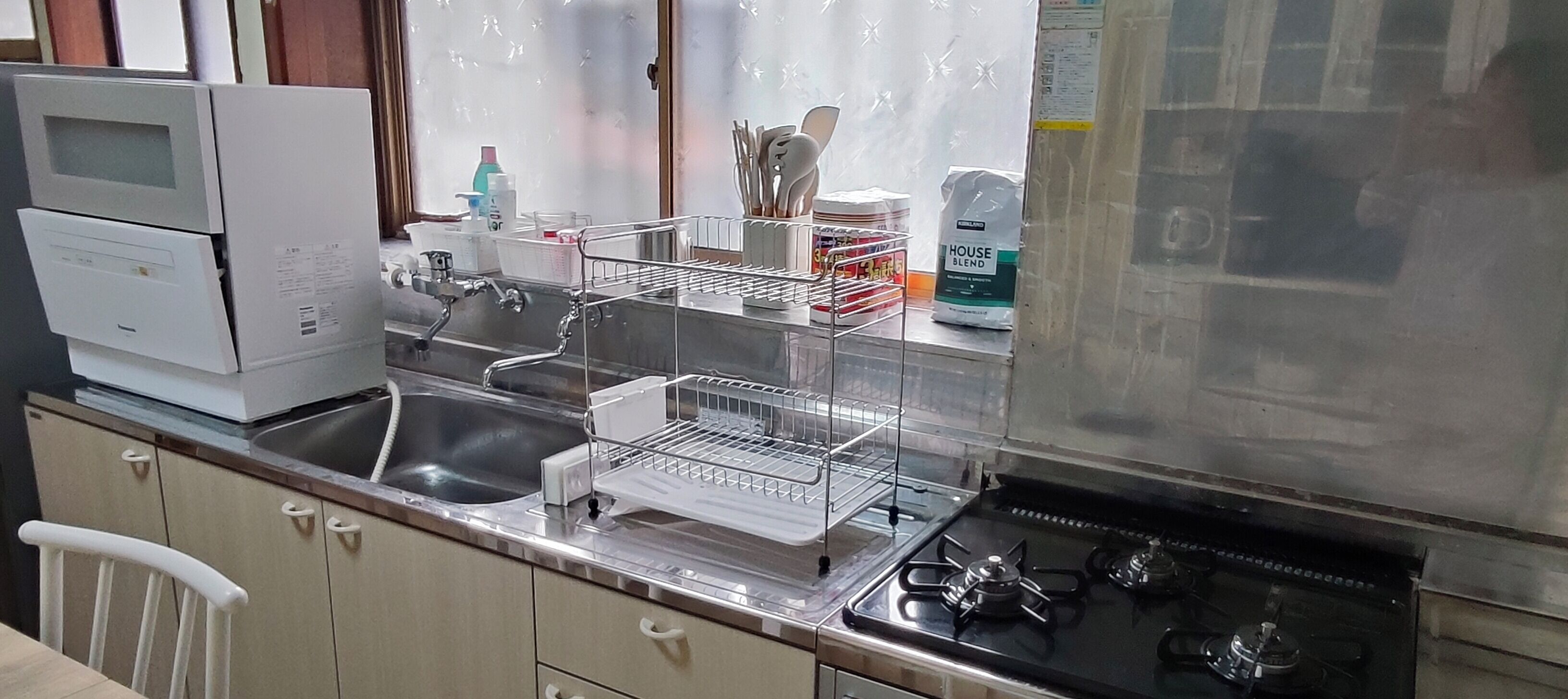 Une cuisine équipée d’un évier en acier inoxydable, d’une cuisinière et d’un four à micro-ondes. On y trouve des placards, une chaise et divers ustensiles de cuisine, comme un égouttoir et un sac de farine.