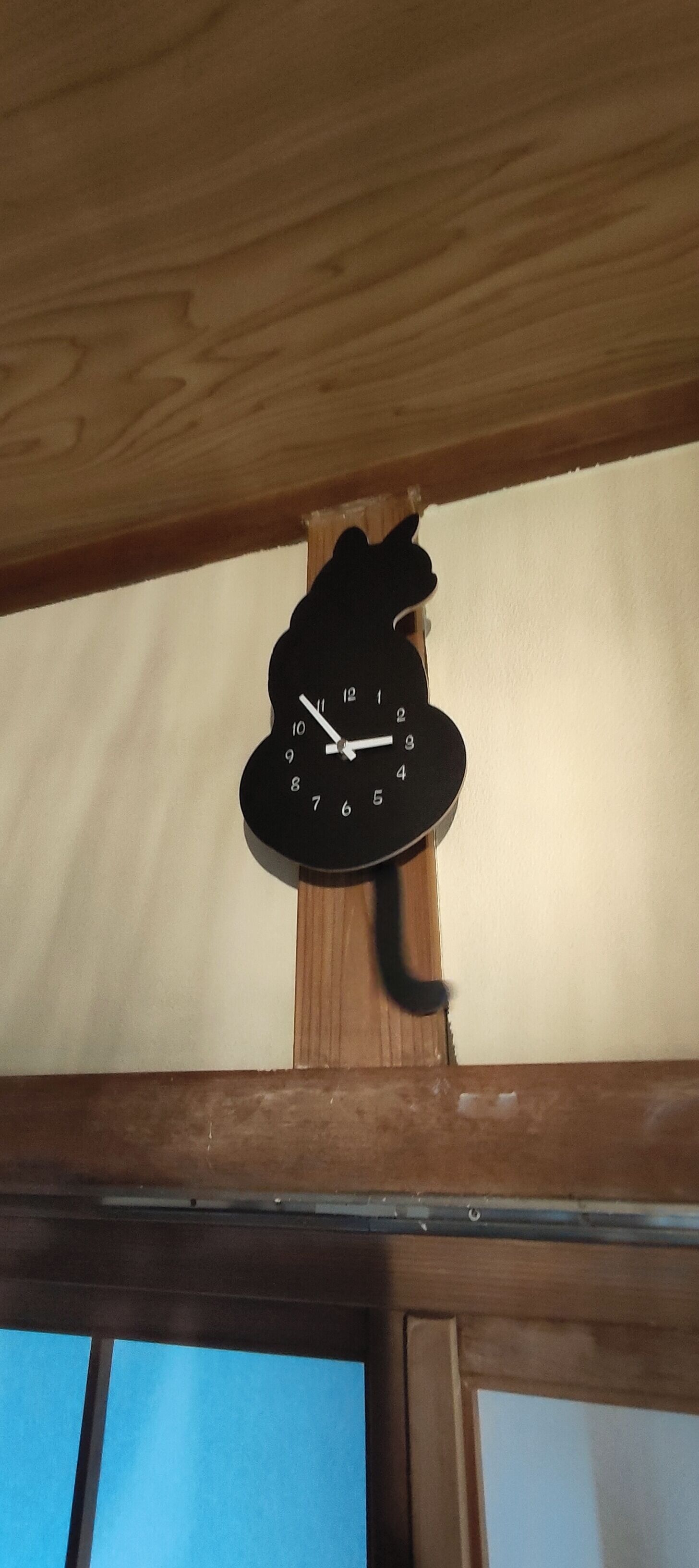 A wall-mounted clock with a cat silhouette design, positioned above a window with a wooden frame.