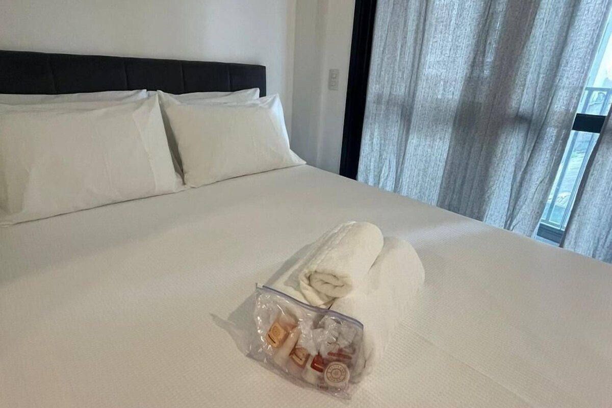 A hotel room with a neatly made bed, a black headboard, an air conditioning unit, and a window with curtains.