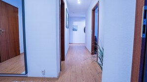 Un couloir étroit avec un plancher en bois, des murs blancs et un miroir sur le côté gauche.