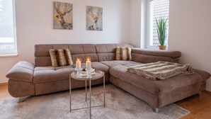 Un salon moderne avec un canapé d’angle, deux tableaux accrochés au mur, une petite table d’appoint avec des bougies et une plante en pot près de la fenêtre.