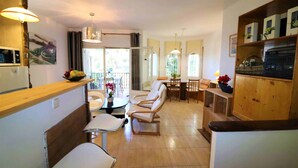 Amplia sala de estar con mesa y sillas de comedor, y una zona de cocina con refrigerador y estanterías de madera.