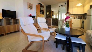 Una sala de estar moderna con una mesa de comedor redonda, dos sillones blancos acolchados y un televisor sobre un soporte de madera.