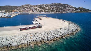Zona costera con un rompeolas rocoso, un puerto deportivo con barcos y una ciudad en una colina al fondo.