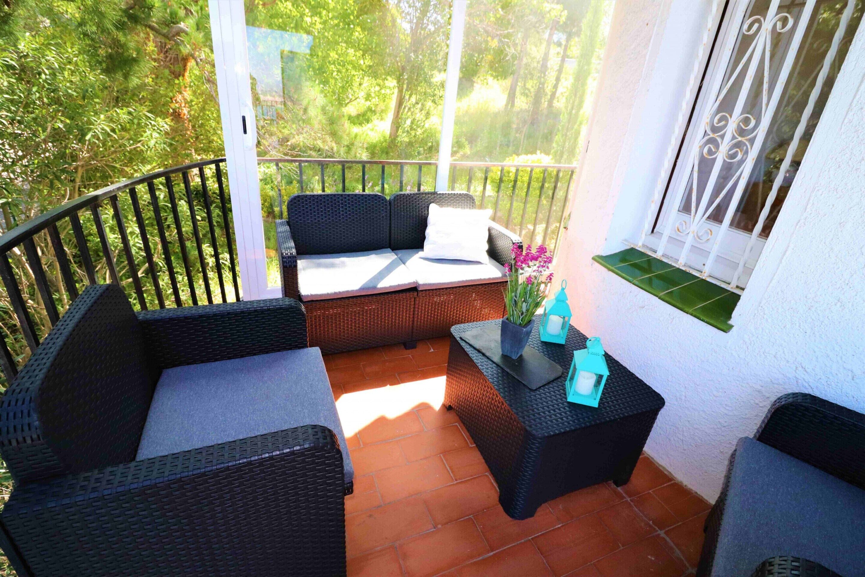 A balcony with wicker furniture, a small table with a vase and lantern, and a window with decorative iron bars.