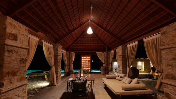 A cozy, rustic room with a stone wall, a wooden ceiling, a sofa, a table, and a chair. - Neem Sarai Sariska (Rajgarh)