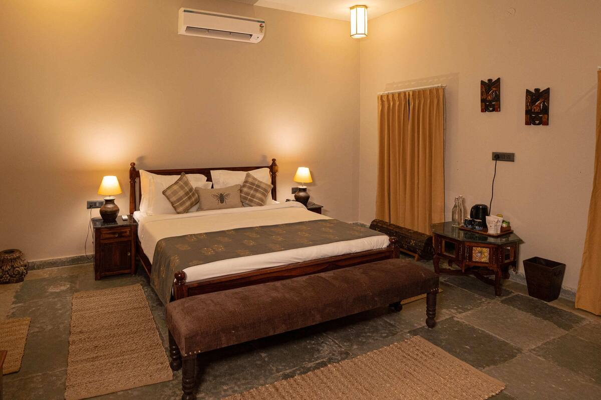A neatly arranged bedroom with a large bed, two bedside tables with lamps, a small table with a kettle, and a trash bin.