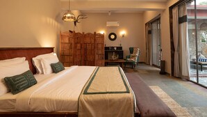 A hotel room with a large bed, a wooden headboard, a decorative wall panel, a seating area with a chair and table, and a view through a glass door. - Neem Sarai Sariska (Rajgarh)