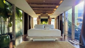 A spacious bedroom with a large bed, wooden ceiling beams, and a view of greenery through large windows. - Kawung Villa Canggu (Canggu)