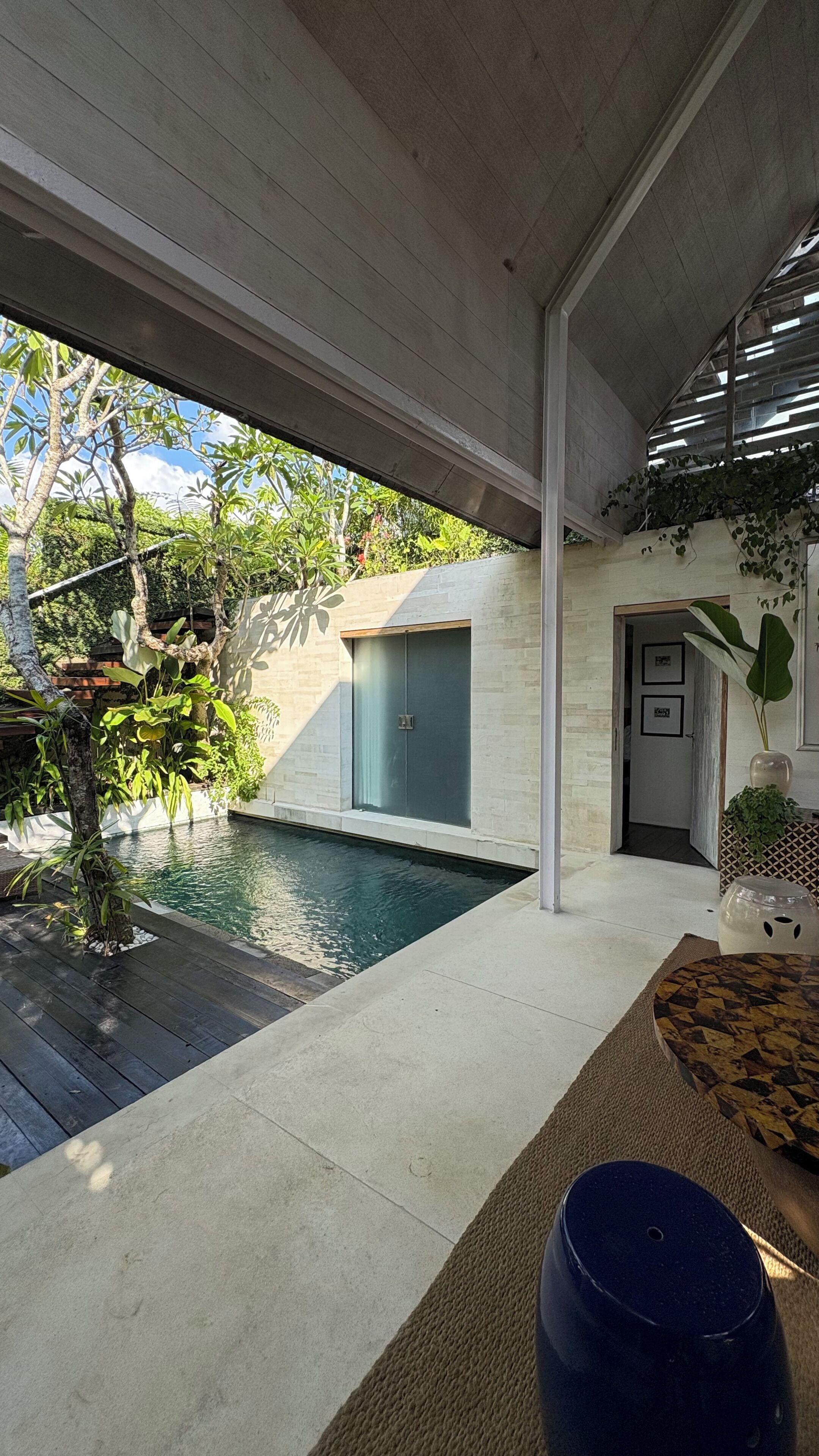 A modern outdoor pool area with a wooden deck, a concrete floor, and a glass door leading to a covered patio.