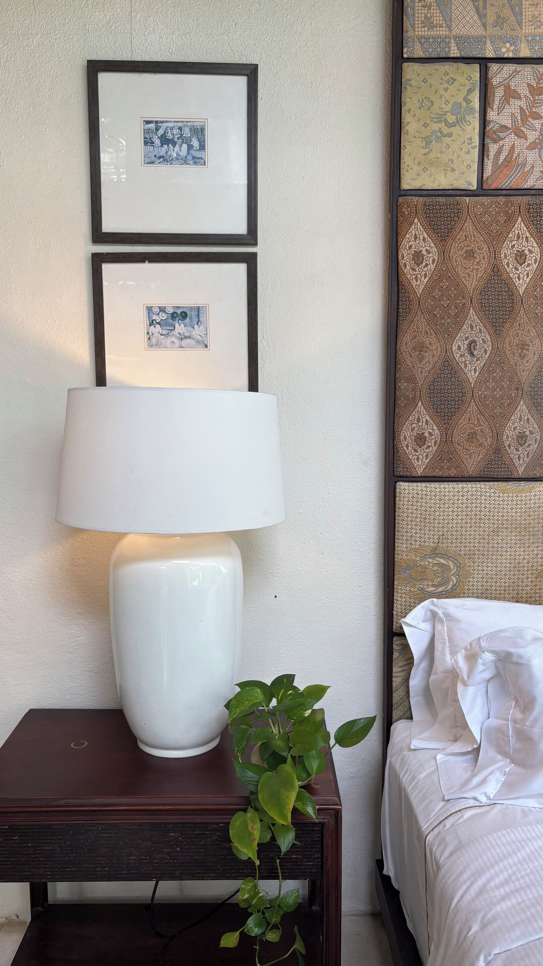 A neatly arranged hotel room with a bedside table, a lamp, a potted plant, and a bed with white linens.
