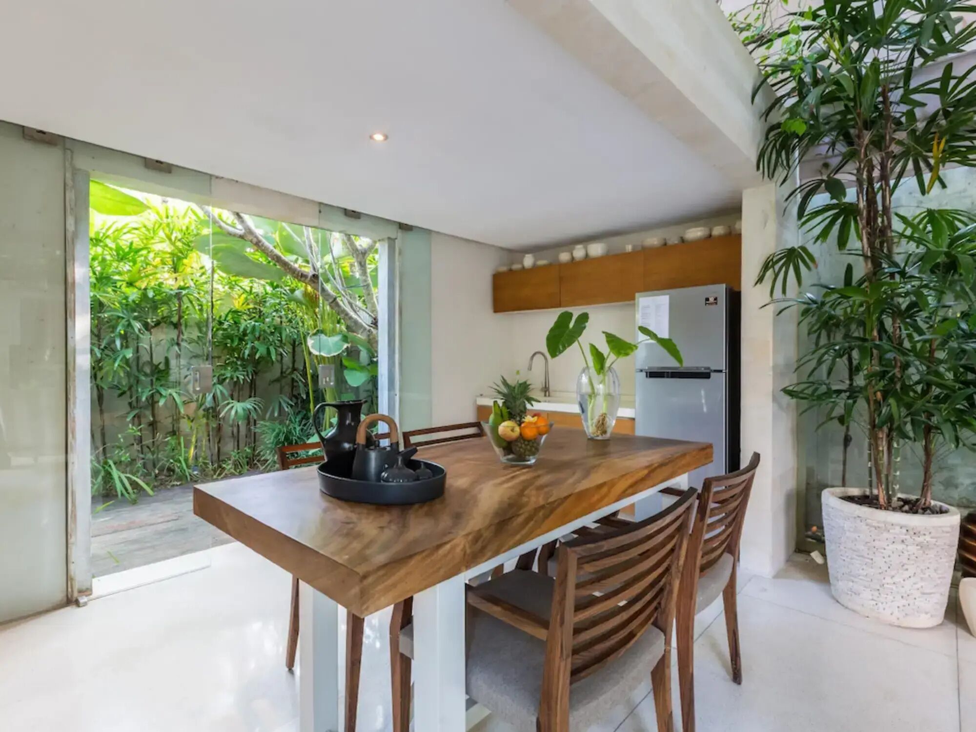 A modern kitchen with a wooden dining table, chairs, and a refrigerator. There is a potted plant on the right and a window with a view of greenery outside.