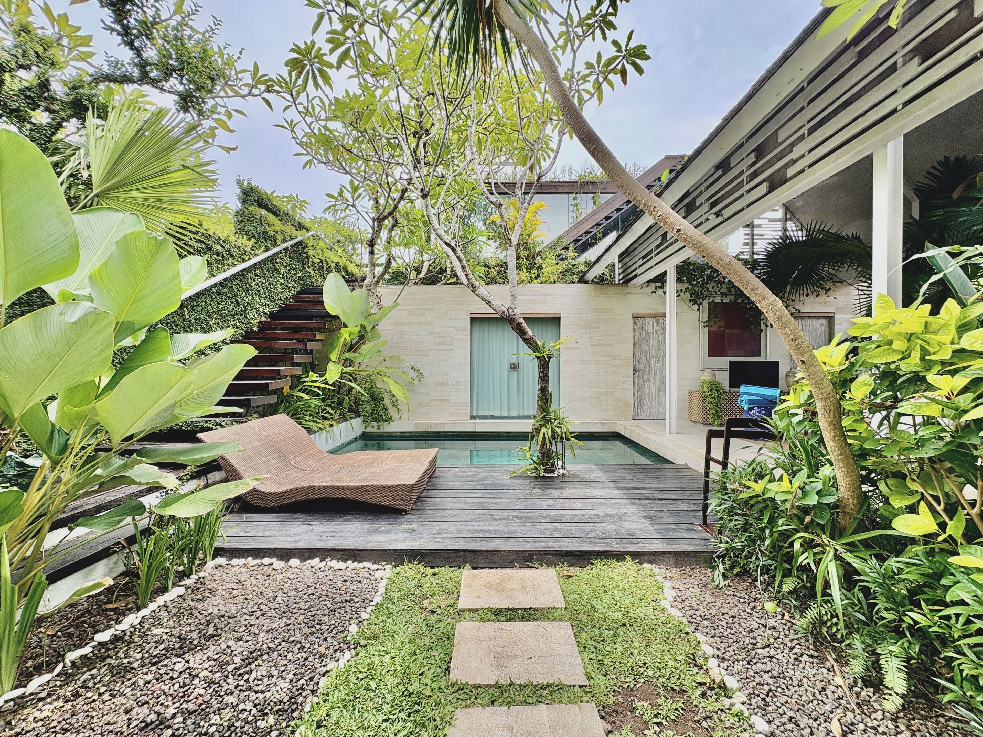 Un área de piscina moderna al aire libre con cubierta de madera, rodeada de abundante vegetación y una escalera que lleva a una terraza cubierta.