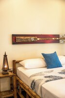 Una cama con ropa de cama blanca y una almohada azul, cabecero de madera, una guitarra colgada en la pared y una mesita con un farol.