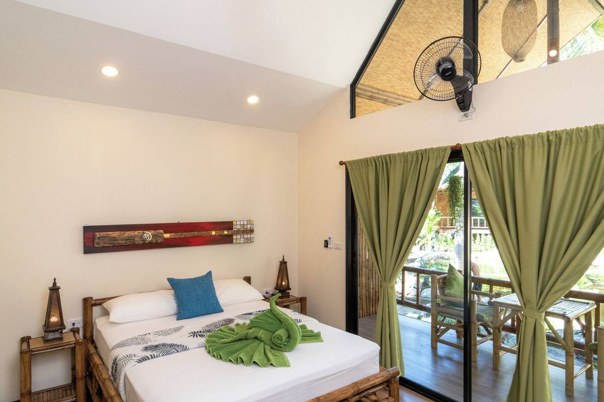 Een slaapkamer met een houten bed, een groot raam met gordijnen, een plafondventilator en een hanglamp.