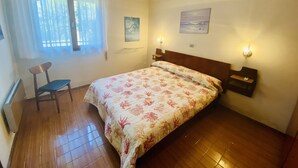 Ein Schlafzimmer mit einem Bett, einem Sessel, einem Fenster mit Vorhängen, einer Wandleuchte und einem Gemälde.
