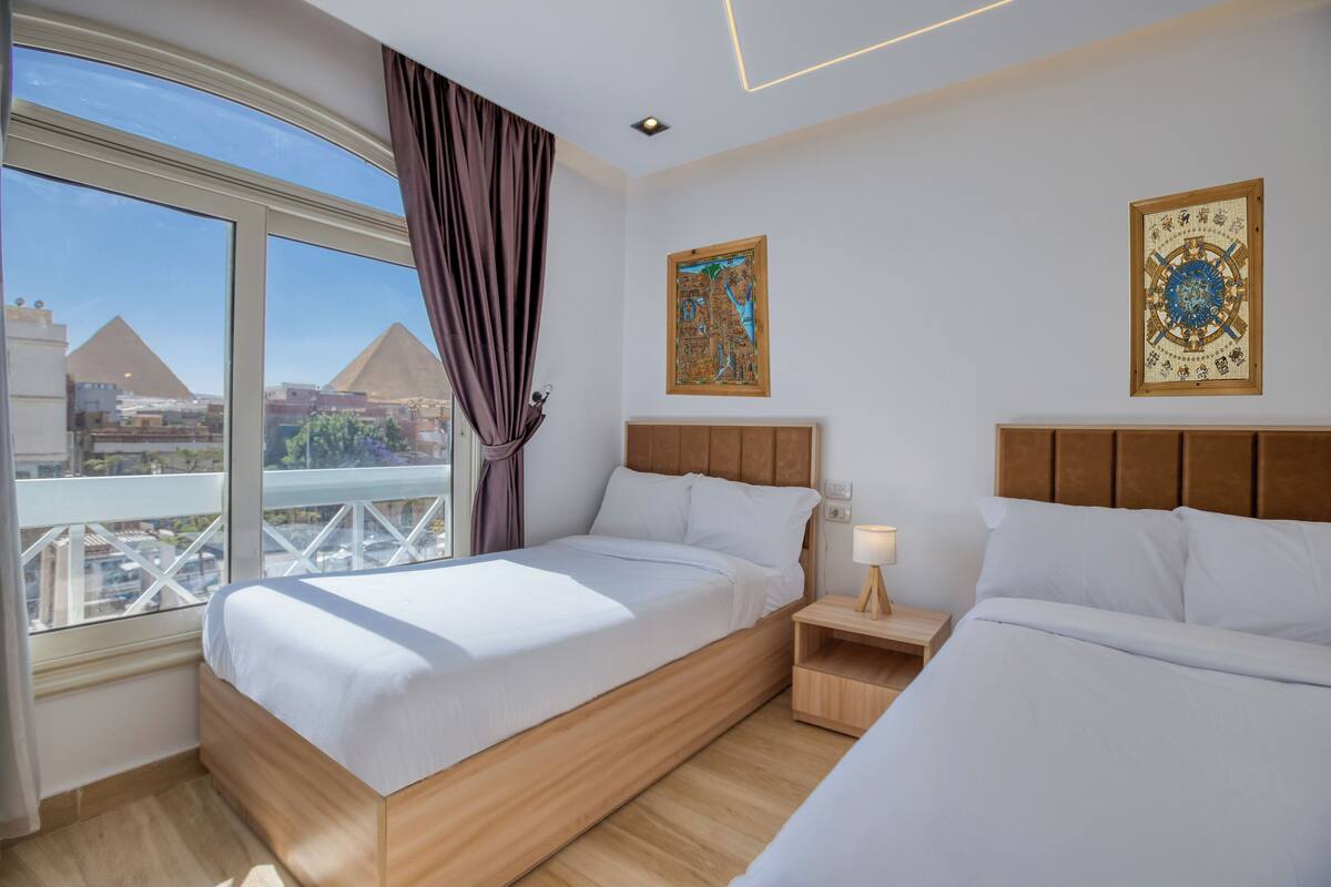A hotel room with two beds, a large window with a view of pyramids, and two wall-mounted artworks.