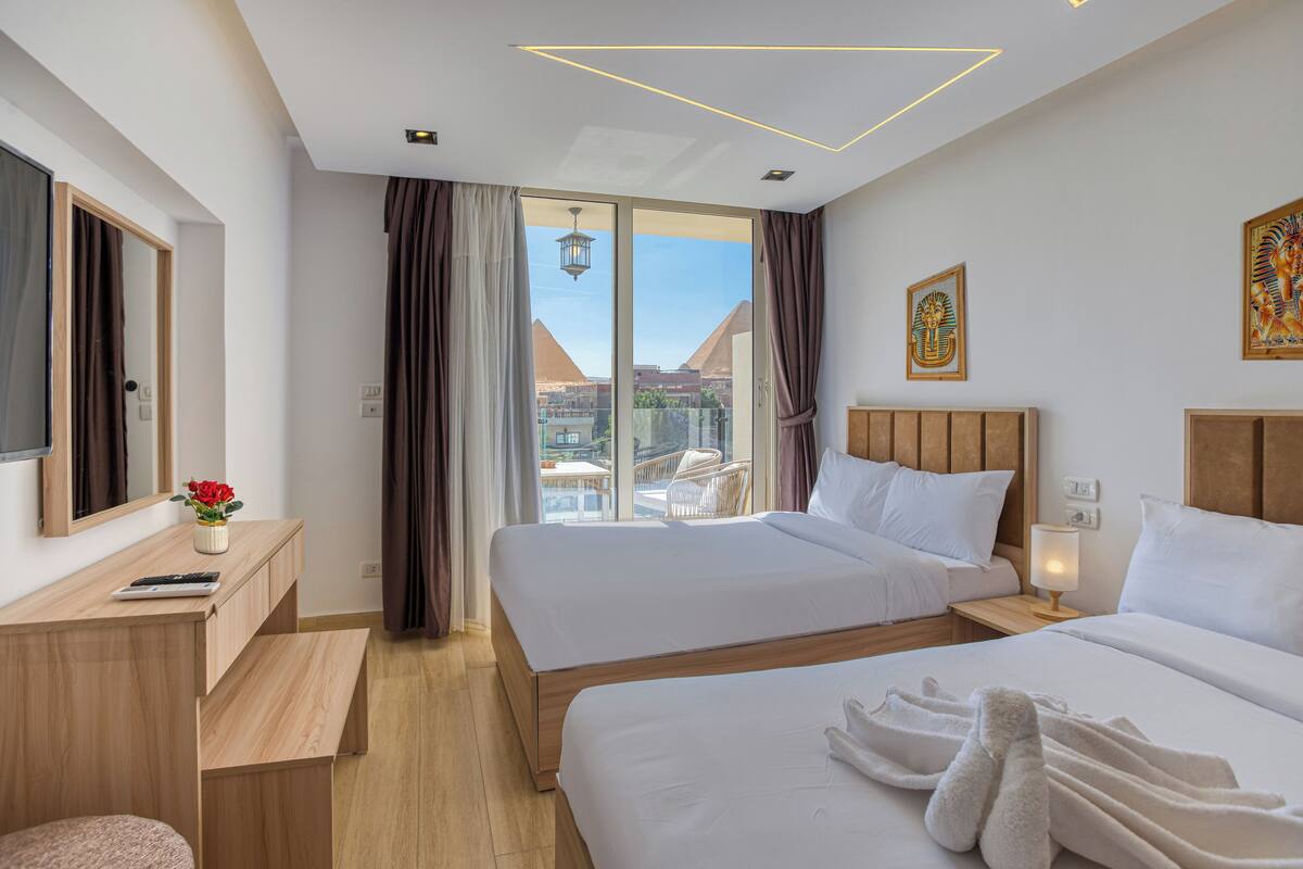 A hotel room with two beds, a desk, a chair, and a view of pyramids.