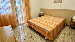 Une chambre d’hôtel avec un lit, un bureau, une chaise, une lampe, un tableau au mur et une fenêtre avec des rideaux.