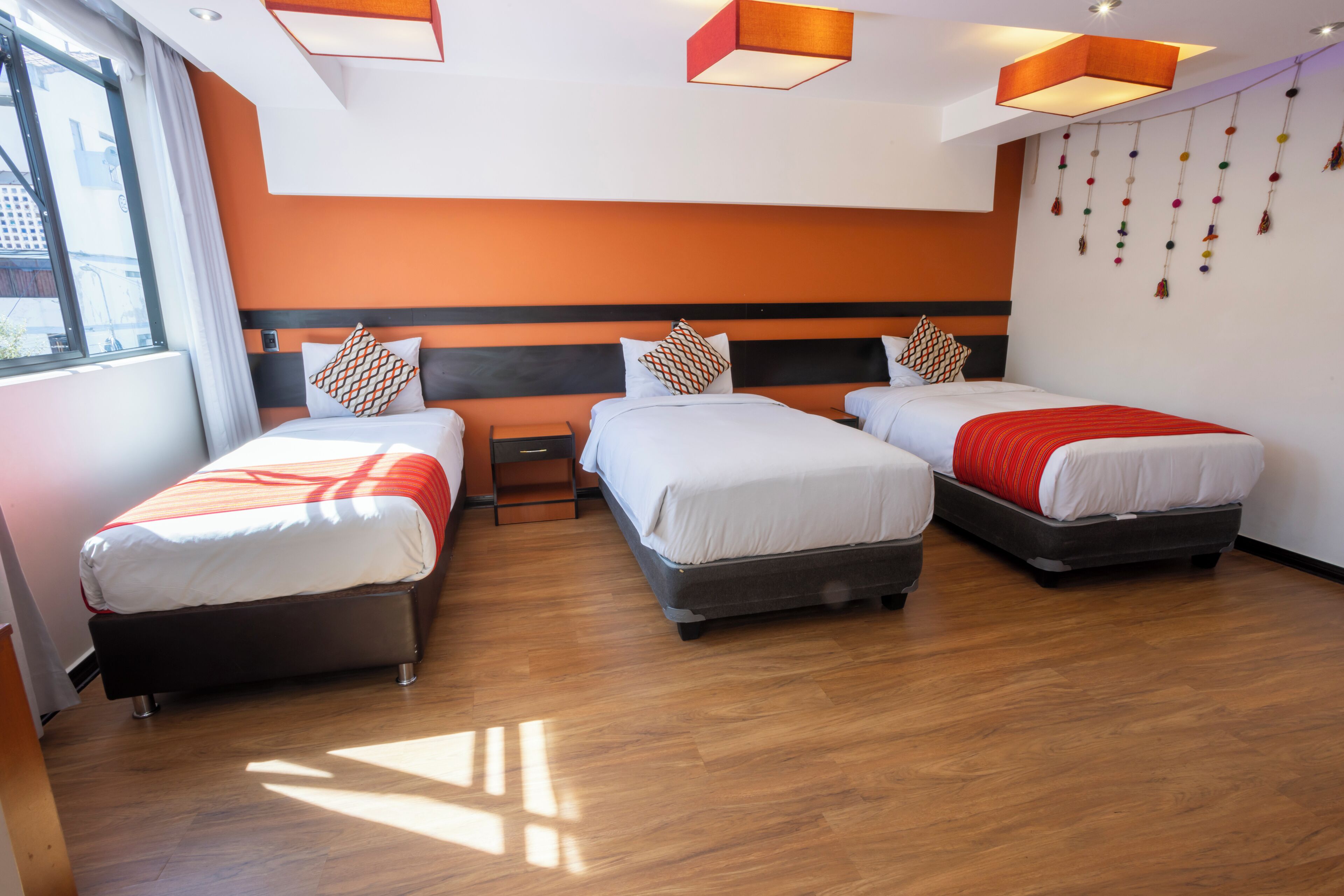 A hotel room with three beds, wooden flooring, and a large window providing natural light.