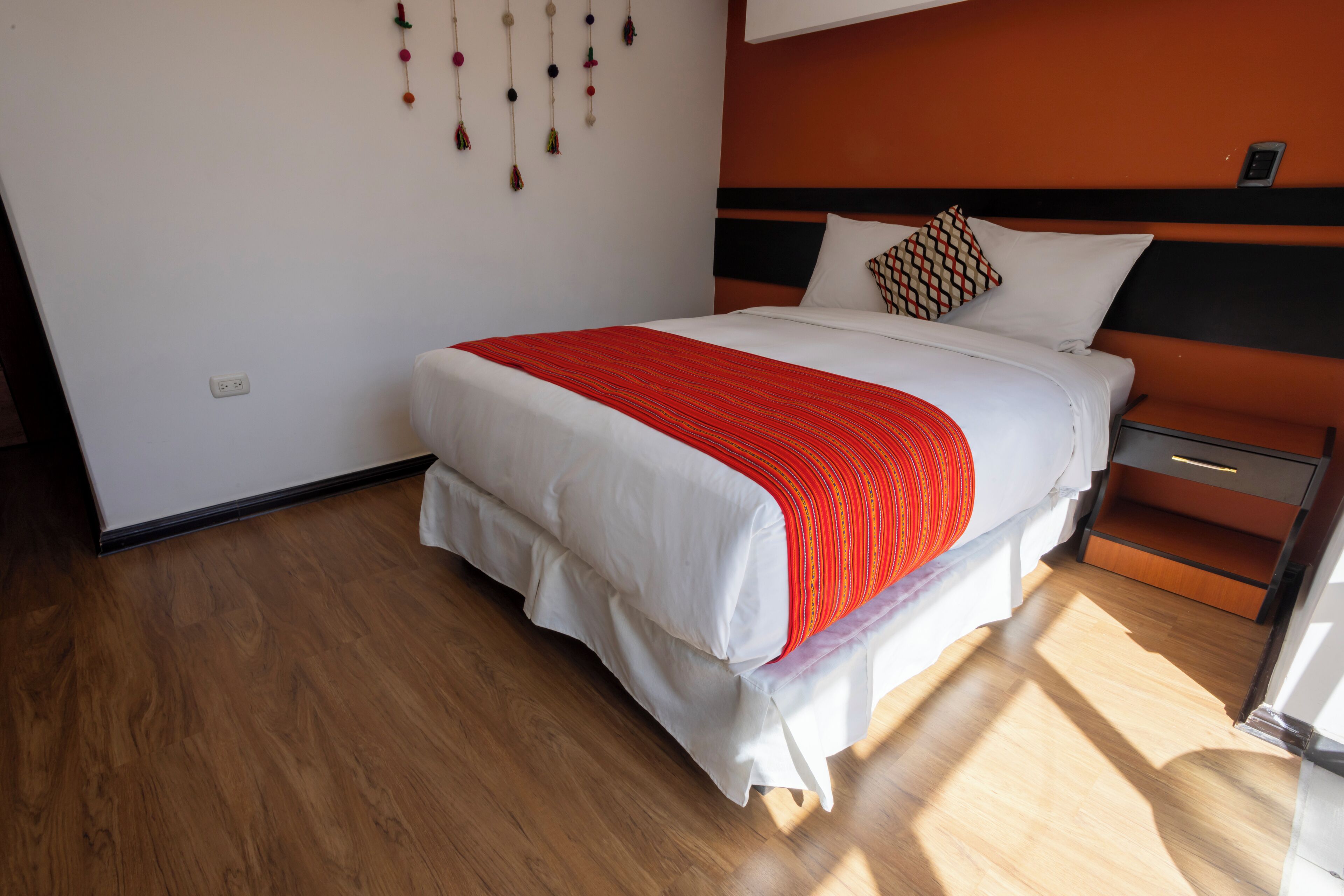 Ein ordentlich bezogenes Bett mit einer rot-weiß gestreiften Decke, Holzboden und einer an der Wand montierten Leuchte.
