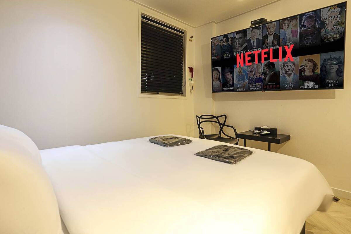 Une chambre d’hôtel avec un grand lit, un téléviseur fixé au mur affichant le logo de Netflix, une petite table avec une télécommande et une chaise.