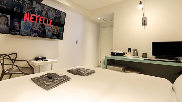 Habitación de hotel con un televisor grande mostrando contenido de Netflix, un escritorio con una computadora, una silla y una cama con un almohadón y un libro.