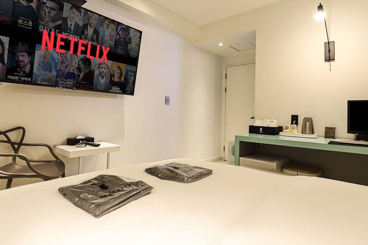 Une chambre d’hôtel dotée d’une grande télévision diffusant du contenu Netflix, d’un bureau avec un ordinateur, d’une chaise et d’un lit avec un oreiller et un livre.