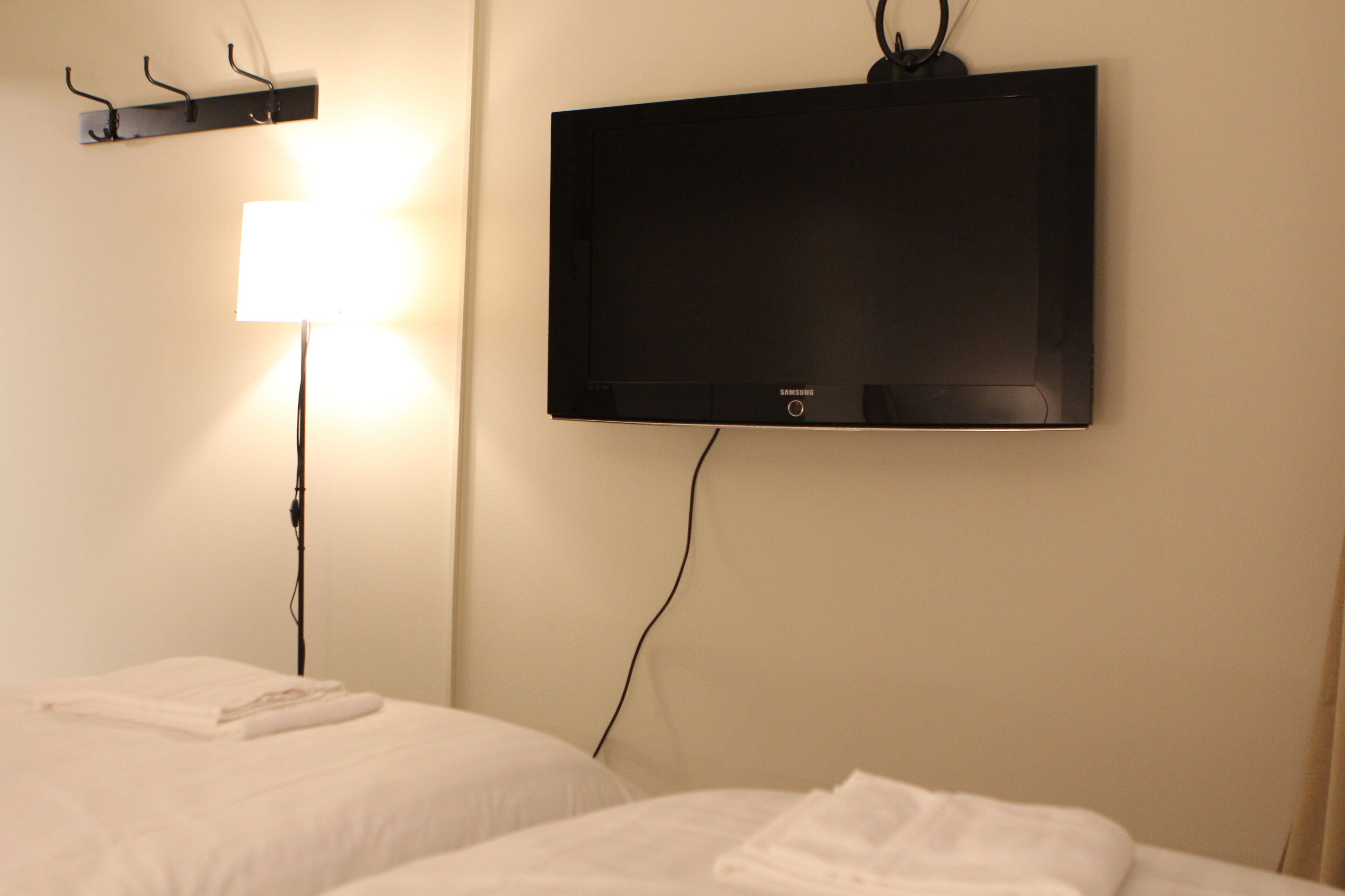 A hotel room with a flat-screen TV mounted on the wall, a bedside table with a lamp, and neatly made beds.