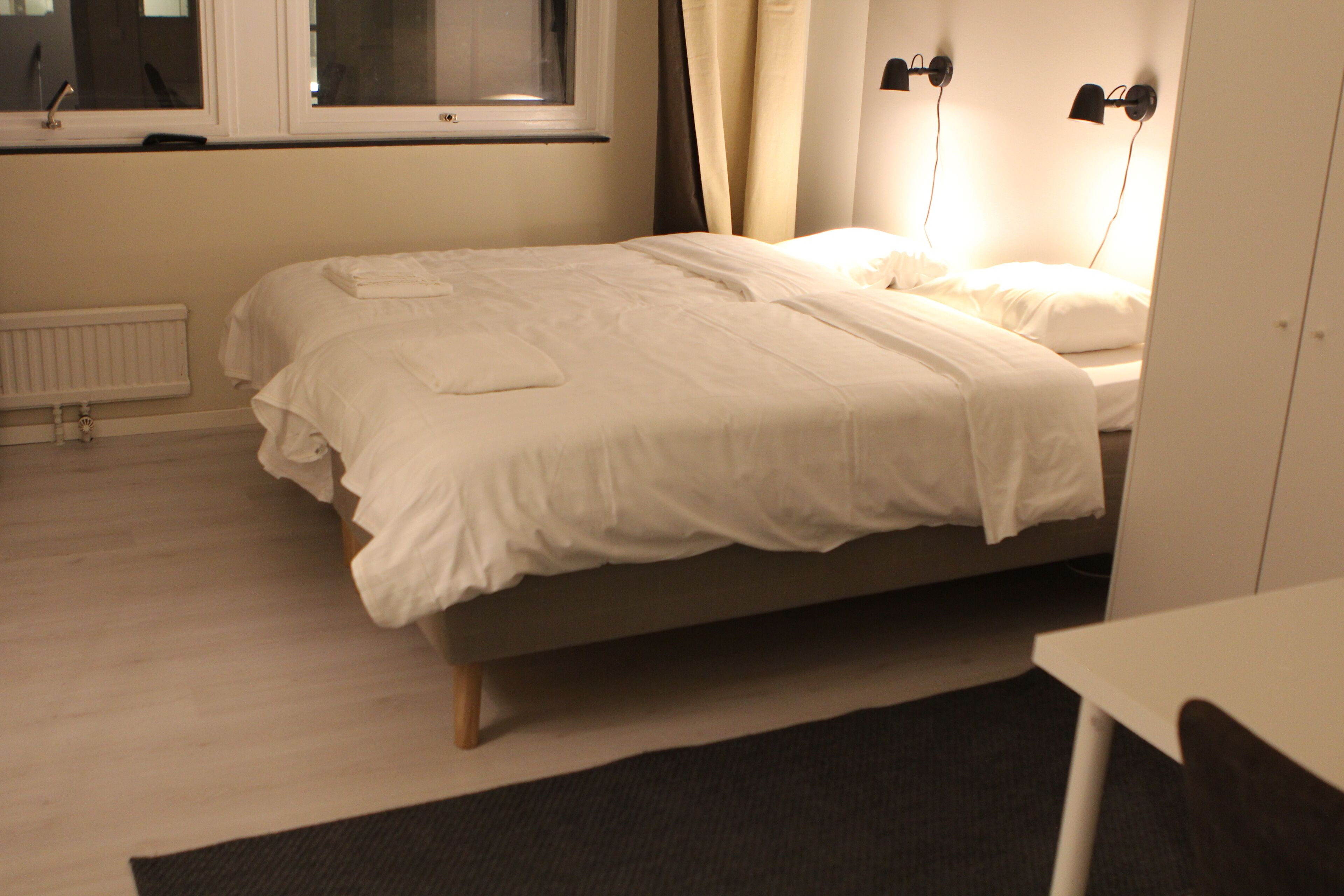 Un letto ben rifatto con coperta bianca e cuscini, un comodino, un termosifone e una finestra con tende.