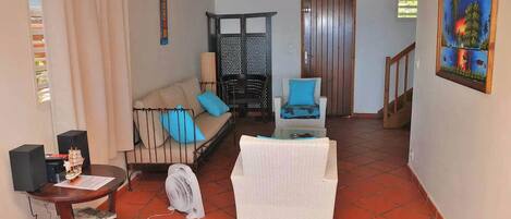 Un salón con sofá, sillón y una mesita con un ventilador. Se ve una escalera que lleva a otra habitación.