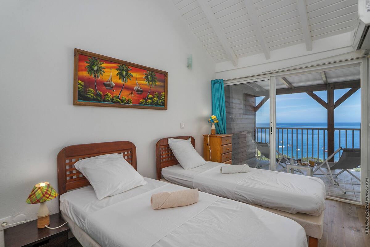 Habitación de hotel con dos camas, un cuadro de paisaje tropical, un aparador de madera y un balcón con vistas al mar.