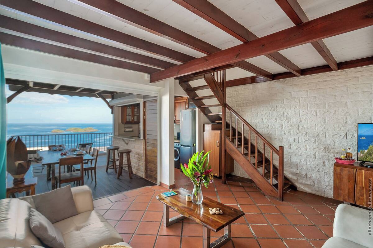 Amplia sala de estar con vista al mar, una escalera de madera y un comedor con mobiliario exterior.