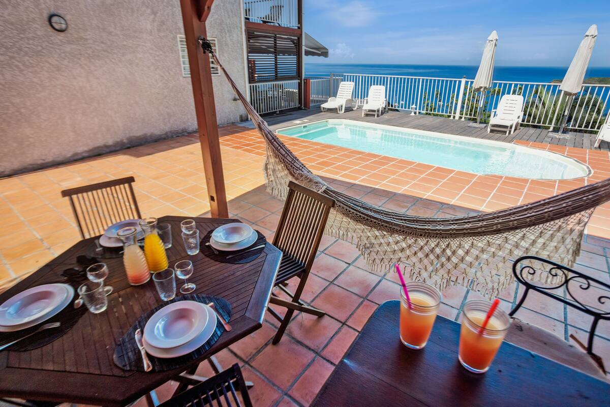 Un área de comedor junto a la piscina, con una mesa preparada para dos, una hamaca y vista al océano.